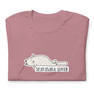 May include: A mauve t-shirt featuring a cartoon cat in a savasana pose with the text "SHAVASANA LOVER".