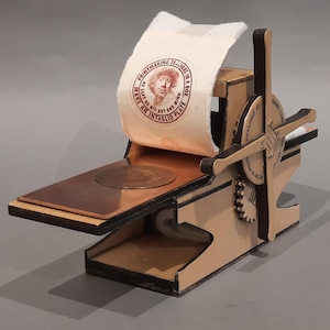 Puede incluir: Una pequeña prensa de impresión de madera con un rollo de papel blanco y una placa redonda de cobre. El rollo de papel tiene un sello circular rojo y blanco con el texto "PRINTMAKING TEACHERS IN A BOX MAKE US HIS ART AND MIND AN INTAGLIO PLATE".