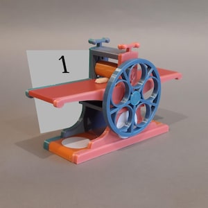 May include: A small, pink and blue 3D printed model of a vintage printing press with a white card that says "1" on it.