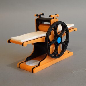 May include: A small, orange and black 3D printed model of a rolling press. The press has a white felt pad and a black roller with a blue accent.