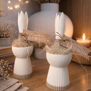 Puede incluir: Dos decoraciones blancas de cerámica con forma de conejito, con base acanalada y ribete marrón. Cada conejito sostiene un manojo de paja y una etiqueta que dice "KLEINIGKEIT". Las decoraciones están sobre una superficie de madera.