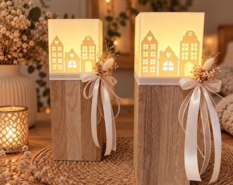 May include: Two rectangular wooden lanterns with illuminated white paper shades featuring a cutout village house design. Each lantern is decorated with a white satin ribbon bow and a small cluster of dried flowers attached to the front.