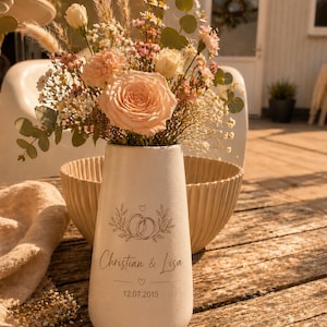 Pode incluir: Um vaso de cerâmica branco mate preenchido com um bouquet de rosas cor de pêssego, ervas secas e pequenas flores brancas. O vaso apresenta um design impresso de dois anéis entrelaçados, os nomes Christian & Lisa e a data 12.07.2015 em tinta preta.