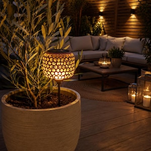 Può includere: Una luce solare da giardino rotonda e scura con un design a nido d'ape traforato, che emette una calda luce gialla. La luce è montata su un sottile paletto nero ed è posizionata all'interno di un grande vaso cilindrico testurizzato chiaro.