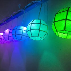 May include: A row of colorful, spherical paper lanterns hanging from a blue pipe. The lanterns are lit with various colors, including orange, purple, blue, and green. Each lantern has a geometric grid pattern.