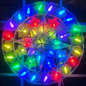 May include: A colorful, illuminated star-shaped Christmas decoration. The design features a central star surrounded by a circular arrangement of smaller stars, each housing a small light. The lights are in various colors, including red, green, blue, and yellow.