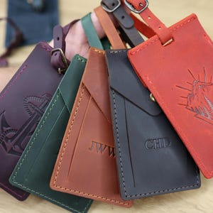 May include: A collection of rectangular leather luggage tags in various colors including red, dark brown, tan, green, and purple. Each tag features a buckle strap and unique embossed designs, such as a mountain scene, anchor, or initials like JWW and CHD.