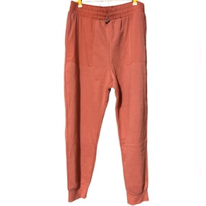 Könnte beinhalten: Korallfarbene Jogginghose mit Kordelzug in der Taille und Seitentaschen. Die Hose ist aus einem weichen Material gefertigt und hat eine lockere Passform. Die Bündchen sind gerippt. Die Hose hängt an einem Kleiderbügel vor weißem Hintergrund.