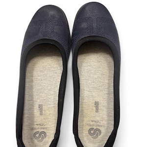 May include: A pair of navy blue Clarks Cloudsteppers ballet flats. The shoes have a textured upper with stitched detailing and a black trim. The interior features a gray cushioned insole with the Clarks Cloudsteppers logo.
