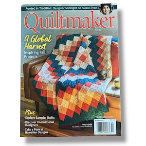 Puede incluir: La portada de la revista "Quiltmaker" presenta un edredón colorido con un diseño geométrico en rojo, naranja, azul y verde, sobre una silla de cuero marrón. El título de la revista es rojo, con el texto "a Global Harvest" y "Inspiring Fall Projects".