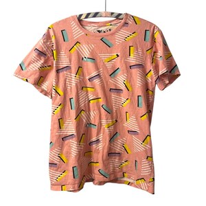 May include: A short-sleeved, peach-colored t-shirt with a repeating geometric pattern. The pattern includes white triangles with parallel lines, and rectangular shapes in yellow, teal, and purple. The shirt is hanging on a hanger.