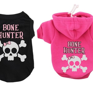 Op de afbeelding: Twee hondenshirts met een schedel en gekruiste beenderen ontwerp en de tekst "BONE HUNTER". Een shirt is zwart met een roze strik op de schedel en de andere is roze met een witte schedel en gekruiste beenderen.