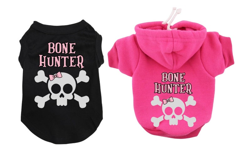 Op de afbeelding: Twee hondenshirts met een schedel en gekruiste beenderen ontwerp en de tekst "BONE HUNTER". Een shirt is zwart met een roze strik op de schedel en de andere is roze met een witte schedel en gekruiste beenderen.