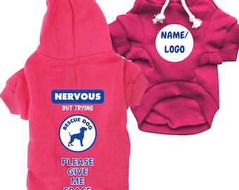 Può includere: Due felpe con cappuccio rosa per cani. Una ha il testo "NERVOUS BUT TRYING RESCUE DOG PLEASE GIVE ME SPACE" e una sagoma di cane. L'altra ha un cerchio bianco con "NAME/LOGO". Entrambe hanno coulisse bianche.