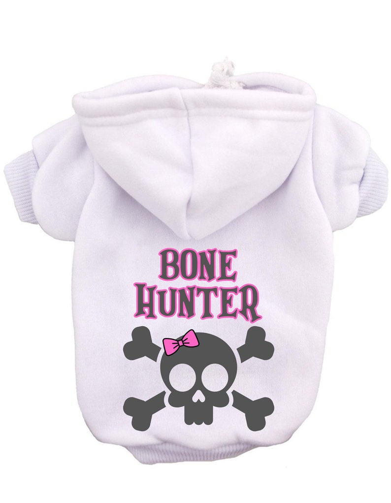 Op de afbeelding: Witte hoodie voor honden met een roze schedel en gekruiste botten afbeelding en de tekst "BONE HUNTER" in roze.