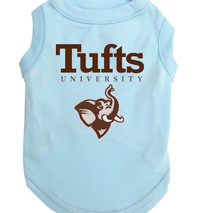 May include: Light blue dog shirt with the text "Tufts UNIVERSITY" in brown block letters. A brown elephant logo is below the text. The shirt is sleeveless and made of a soft material.