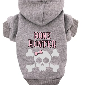 Op de afbeelding: Grijze hoodie voor een huisdier met een witte schedel en gekruiste beenderen afbeelding en de tekst "BONE HUNTER" in roze letters.