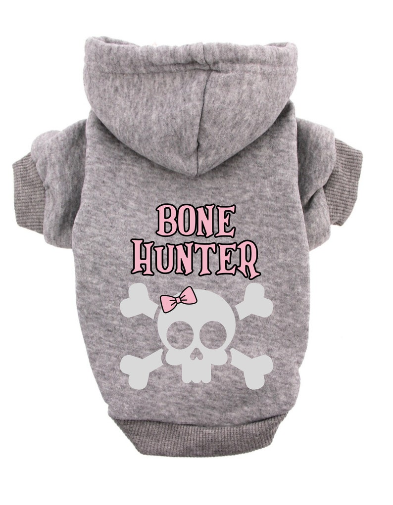 Op de afbeelding: Grijze hoodie voor een huisdier met een witte schedel en gekruiste beenderen afbeelding en de tekst "BONE HUNTER" in roze letters.