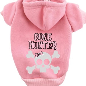 Op de afbeelding: Roze hondenhoodie met een witte schedel en gekruiste botten afbeelding en de tekst "BONE HUNTER" in zwart.