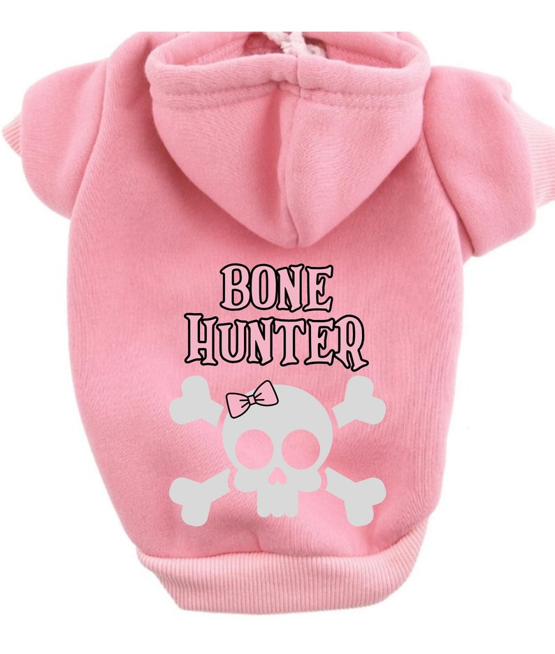 Op de afbeelding: Roze hondenhoodie met een witte schedel en gekruiste botten afbeelding en de tekst "BONE HUNTER" in zwart.
