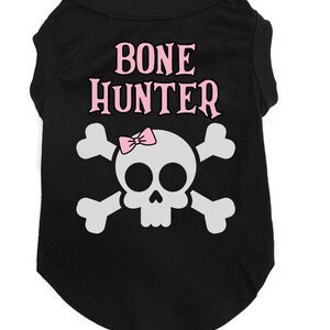 Op de afbeelding: Zwart honden T-shirt met een wit schedel en gekruiste beenderen ontwerp. De schedel heeft een roze strik en de tekst "BONE HUNTER" in roze letters.