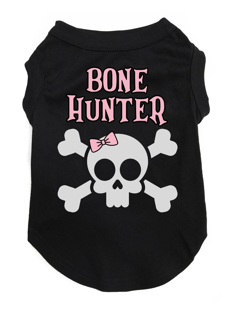 Op de afbeelding: Zwart honden T-shirt met een wit schedel en gekruiste beenderen ontwerp. De schedel heeft een roze strik en de tekst "BONE HUNTER" in roze letters.