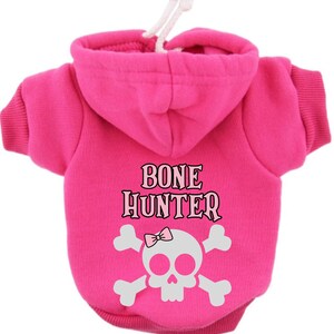 Op de afbeelding: Roze hondenhoodie met een wit schedel- en gekruiste beenderenontwerp en de tekst "BONE HUNTER" op de rug.