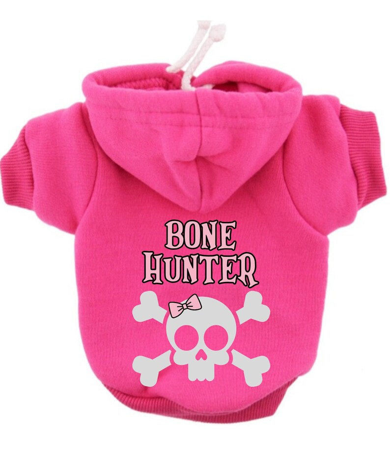 Op de afbeelding: Roze hondenhoodie met een wit schedel- en gekruiste beenderenontwerp en de tekst "BONE HUNTER" op de rug.