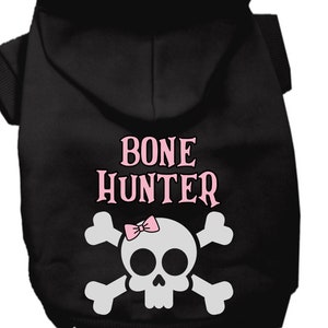 Op de afbeelding: Zwarte hondenhoodie met een witte schedel en gekruiste beenderen afbeelding en de tekst "BONE HUNTER" in roze letters. Een roze strik zit bovenop de schedel.