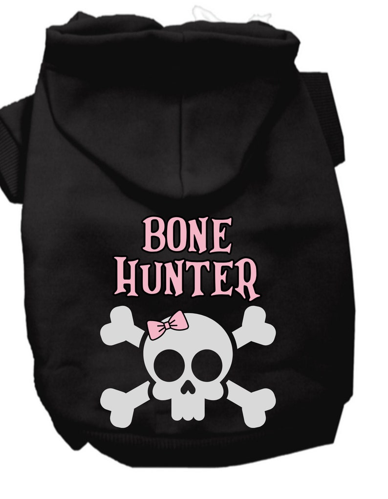 Op de afbeelding: Zwarte hondenhoodie met een witte schedel en gekruiste beenderen afbeelding en de tekst "BONE HUNTER" in roze letters. Een roze strik zit bovenop de schedel.
