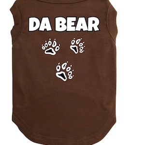 May include: A brown dog t-shirt with the text "DA BEAR" in white block letters. Below the text are three white paw print designs. The shirt is made of a soft material and has a rounded neckline.