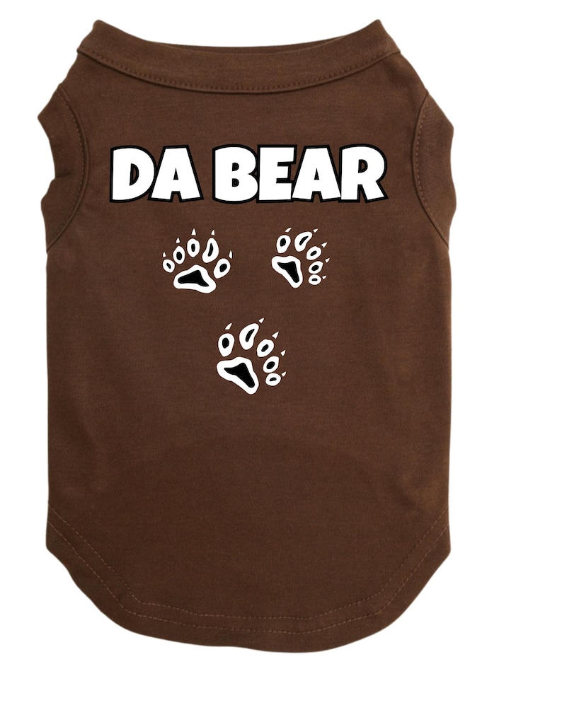 May include: A brown dog t-shirt with the text "DA BEAR" in white block letters. Below the text are three white paw print designs. The shirt is made of a soft material and has a rounded neckline.