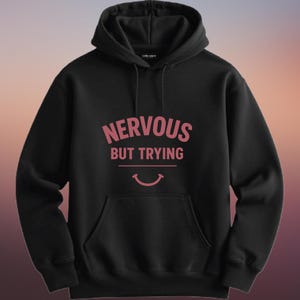 May include: Black hooded sweatshirt with the text "NERVOUS BUT TRYING" in pink letters above a smiling mouth graphic. The sweatshirt has a front pocket and a drawstring hood.