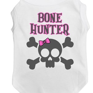 Op de afbeelding: Witte hondenshirt met een roze schedel en gekruiste beenderen ontwerp. De tekst "BONE HUNTER" is in roze letters op het shirt gedrukt.