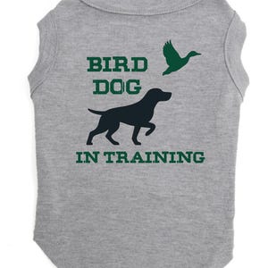 May include: A gray dog shirt with the words "BIRD DOG IN TRAINING" in green block letters. A black silhouette of a dog and a green silhouette of a flying duck are also on the shirt.