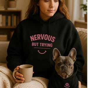 May include: Black hoodie with the text "NERVOUS BUT TRYING" in pink. A person is holding a beige mug. A French Bulldog wearing a matching black hoodie with a paw print and smiling face graphic.