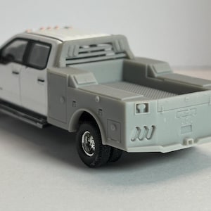 May include: A white and gray toy pickup truck with a flatbed and dual rear wheels. The truck has a detailed design, including a cab, bed, and storage compartments. The tires are black, and the truck appears to be a model.
