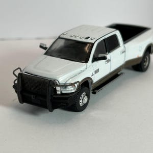 May include: A white and brown pickup truck model with a black front grill guard. The truck has a white cab and a brown lower body. The tires are black, and the truck bed is open. The model has a detailed design.