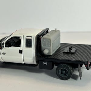 May include: A white pickup truck model with a black flatbed and a gray tank. The truck has black wheels and a detailed interior. The flatbed features a black metal surface and a hitch.
