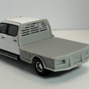 May include: White pickup truck with a flatbed and a metal grate behind the cab. The truck has a gray bed and a black bumper.