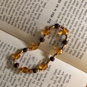 Könnte beinhalten: Eine braune und bernsteinfarbene Perlenkette mit goldener Akzent. Die Kette liegt auf einem Buch mit deutschem Text.