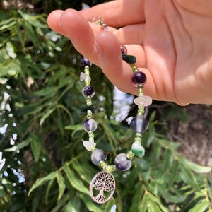 May include: A necklace with a silver tree of life charm, featuring green, purple, and clear beads.