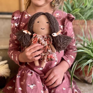 May include: A handmade cloth doll with brown yarn hair styled in pigtails, adorned with small pink ribbons. The doll has a floral print dress and a simple, smiling face. The doll is being held by a person wearing a matching floral dress.