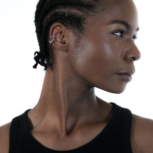 May include: A woman with short black braided hair wearing a black tank top. She is looking to the right side of the frame. She has a silver hoop earring in her left ear.