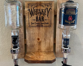 May include: A wooden wall mounted whiskey bar with two clear glass liquor bottles and metal spouts. The sign reads "Welcome Whiskey Bar Good Friends. Good Drinks. Great Times".