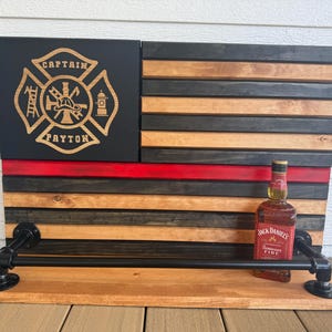 May include: Wooden American flag-themed shelf with a black, red, and natural wood finish. The top left corner features a firefighter emblem with the name "CAPTAIN PAYTON". A bottle of Jack Daniel's Fire whiskey sits on the shelf.