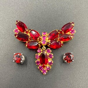 Puede incluir: Un broche de pedrería roja y rosa con un colgante. El broche tiene forma de lazo y tiene un grupo de pedrería roja en el centro. El colgante también está hecho de pedrería roja y tiene forma de lágrima. El broche viene con dos pendientes a juego.
