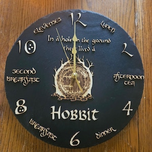 Peut inclure: Une horloge ronde en ardoise noire avec des aiguilles dorées, sur le thème du Hobbit. Le cadran de l'horloge comporte le mot "Hobbit" et la phrase "In a hole in the ground they lived a." L'horloge indique également les heures des repas des Hobbits.