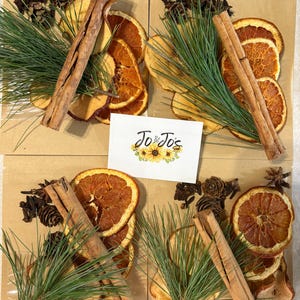 May include: Four brown paper pouches containing dried orange slices, apple slices, pine needles, cinnamon sticks, pinecones, and cloves. A white card with the text Jo & Jo's and a sunflower illustration is placed in the center of the four pouches.