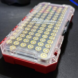 May include: A red and clear plastic ammunition case filled with brass-colored bullets. The case has the "Milwaukee" logo on the top and is sitting on a black surface.